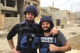 Journalist Matt Brown and cameraman Aaron Hollett stand in an empty street in combat gear front of unfinished buildings.
