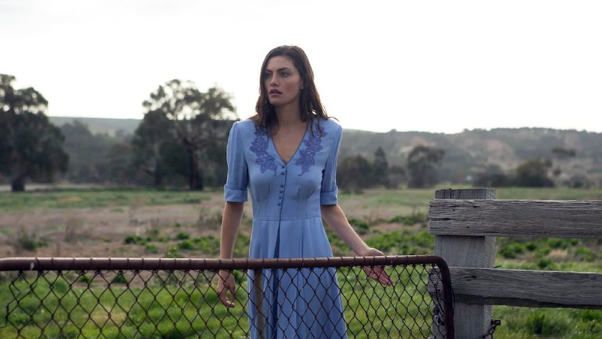 A beautiful young woman stands at a paddock gate in rural Australia.