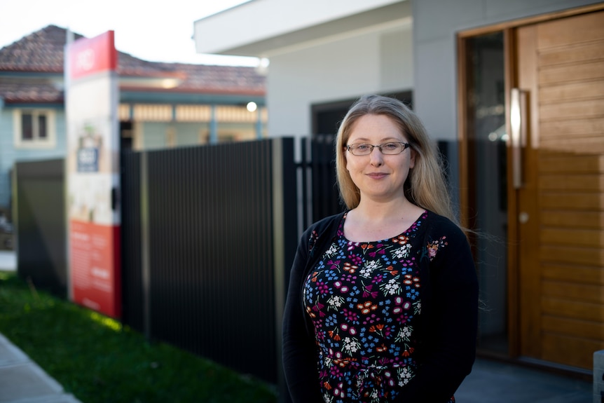 Blond haired woman with glasses smiles at the camera. Behind her is a house with a sold sign