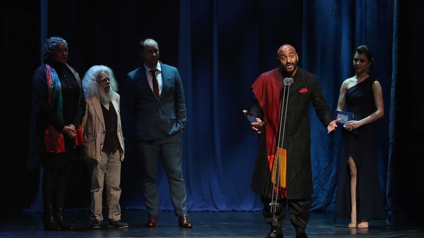 Five people stand on stage, one man stands at the microphone.