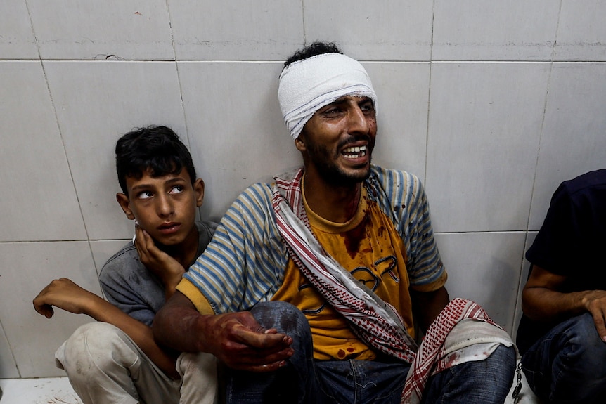 A man with his head bandaged and a pained expression sits on a hospital floor next to a young boy