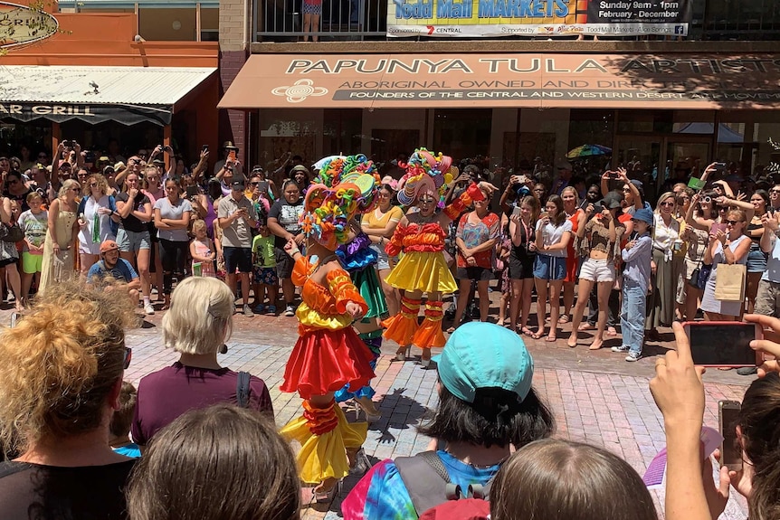 Three performers in yellow, orange and red 'drag' perform on a mall in front of a crowd of people.