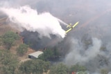 Water emerges from the back of a yellow plane, which flies over burning bushland.