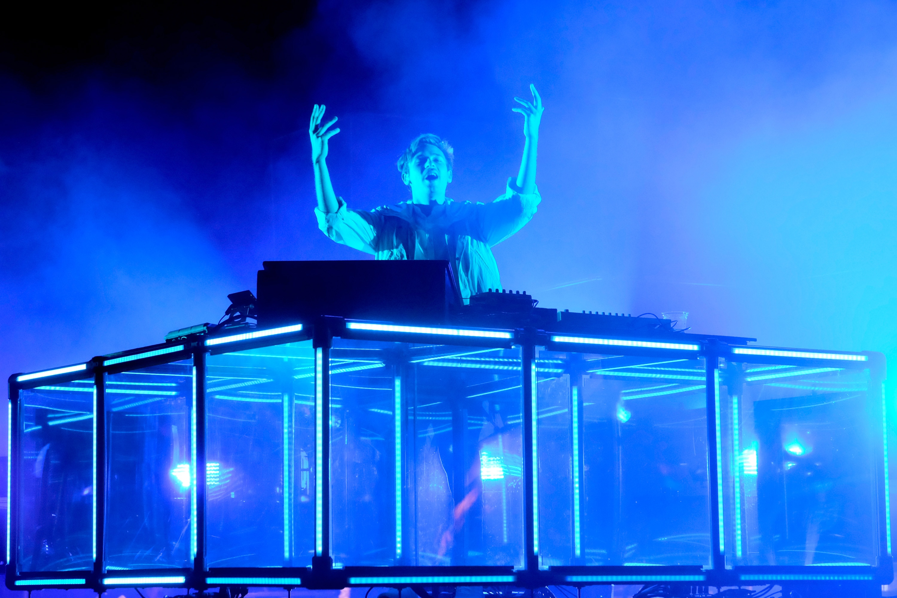 Flume stands behind a glass DJ booth at a festival with his arms raised in the air. He is bathed in blue light.