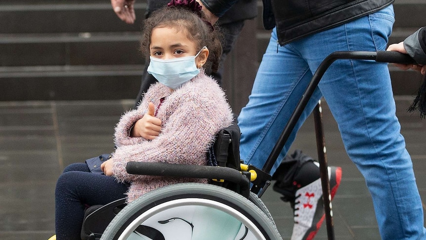 A young girl in a wheelchair wearing a mask and winter clothes gives a thumbs up as she's pushed.