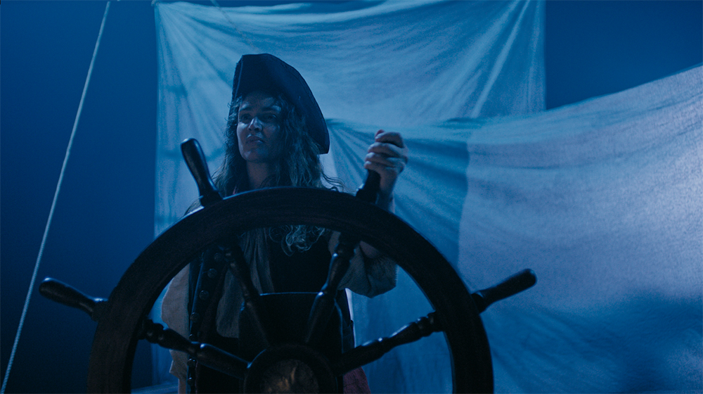 A woman with long dark curly hair wears black pirate holds steering wheel of colonial boat on a dark blue-hued night.