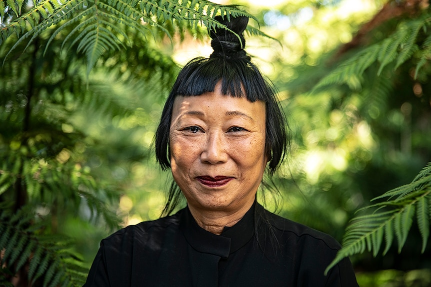 A woman with black hair tied up in top knot and Mao collar style jacket smiles in lush green outdoor area surrounded by ferns.