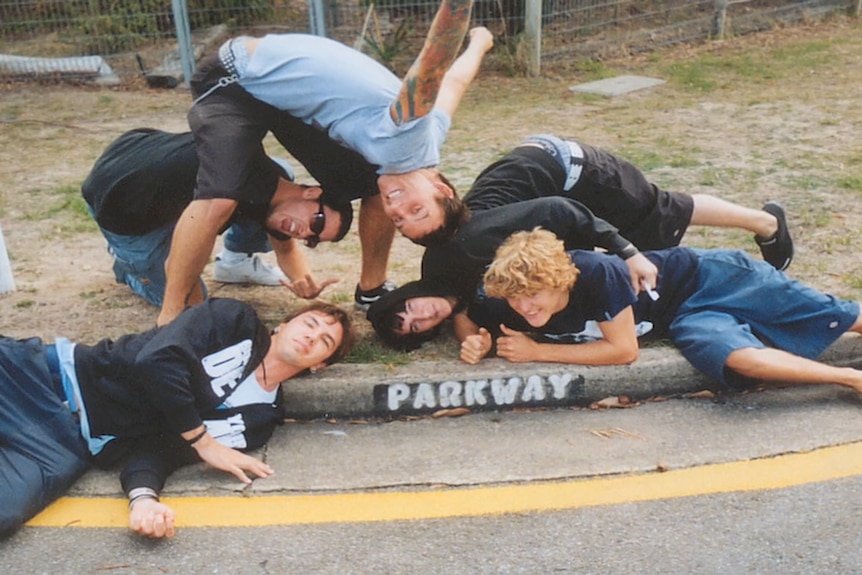 A group of five teenage boys point to a street gutter that reads 'Parkway Drive' in white paint