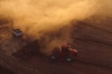 an aerial shot of a tractor pulling a crop seeder through dusty paddock. it's clear from the thick dust that the soil is dry.