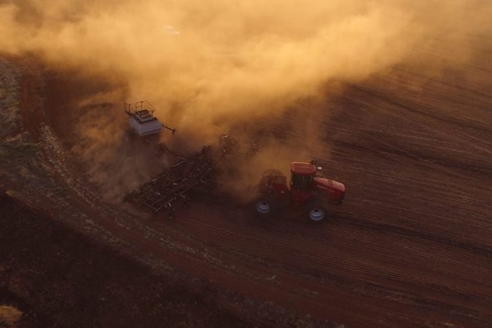 an aerial shot of a tractor pulling a crop seeder through dusty paddock. it's clear from the thick dust that the soil is dry.