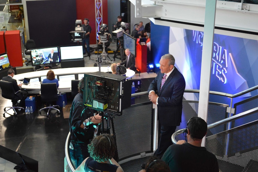 Ian Henderson stands on a staircase and speaks into a camera, in front of an election broadcast set.