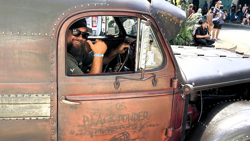A man behind the wheel of a vintage dark coloured hot rod during a car street parade.