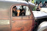 A man behind the wheel of a vintage dark coloured hot rod during a car street parade.