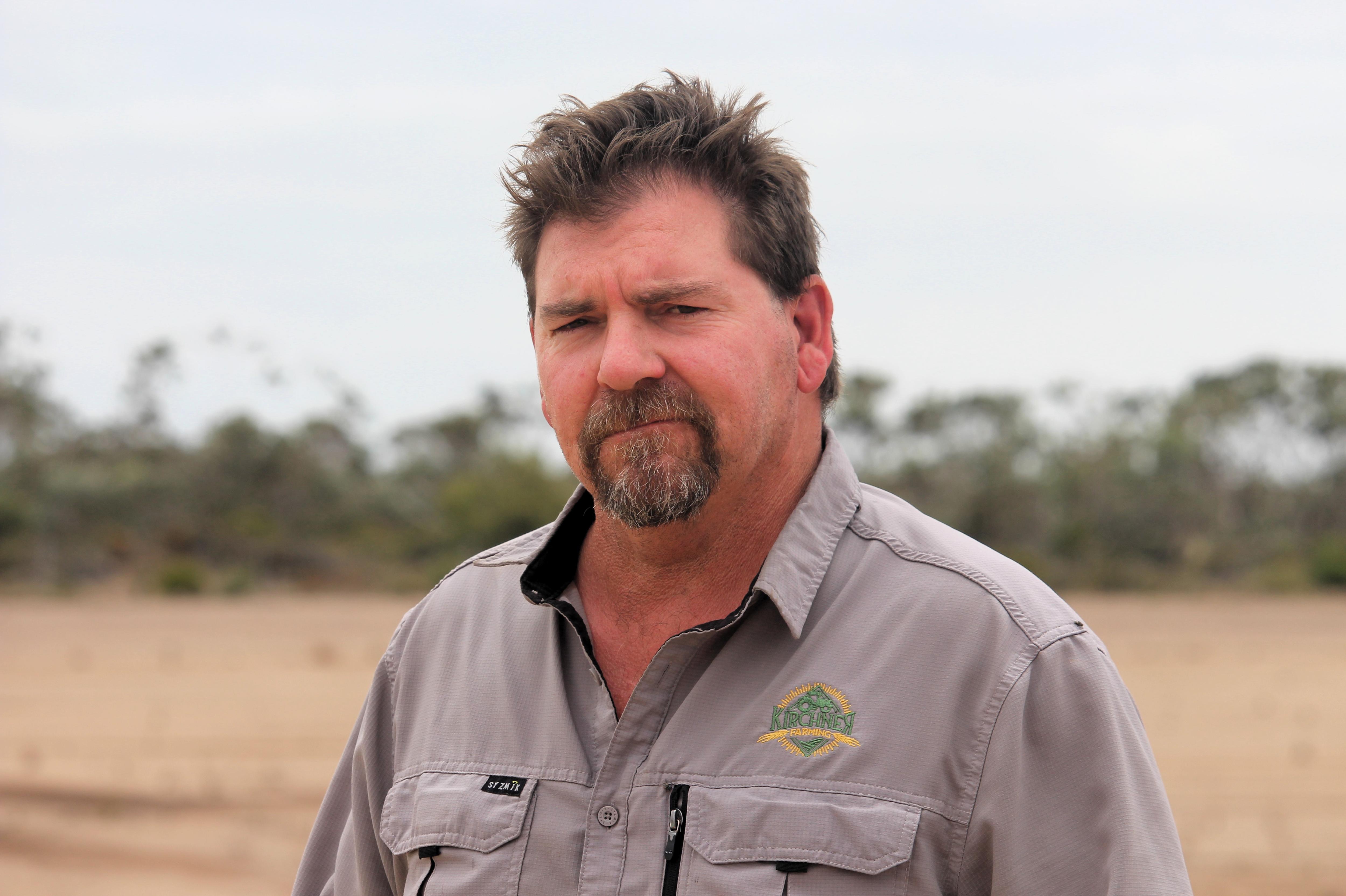 He has brown hair and a beard and stands near gums and a dry paddock