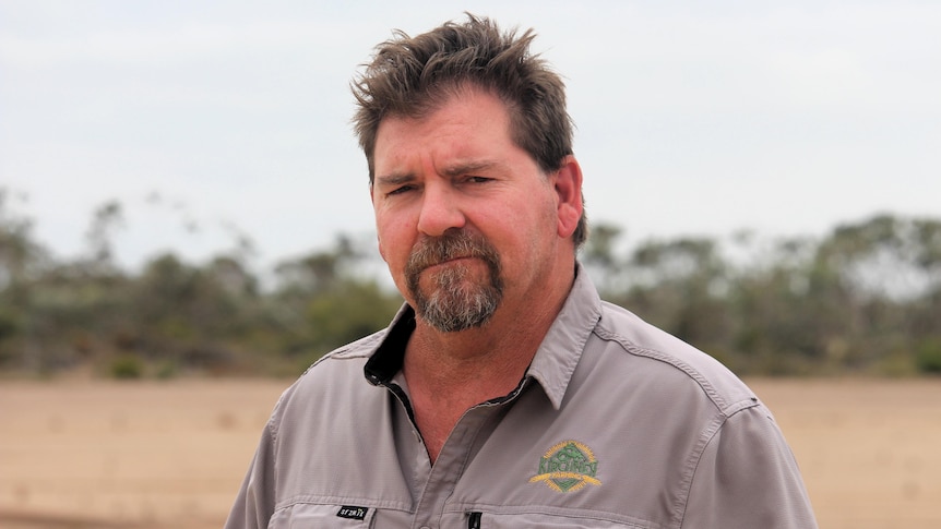 He has brown hair and a beard and stands near gums and a dry paddock