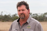 He has brown hair and a beard and stands near gums and a dry paddock
