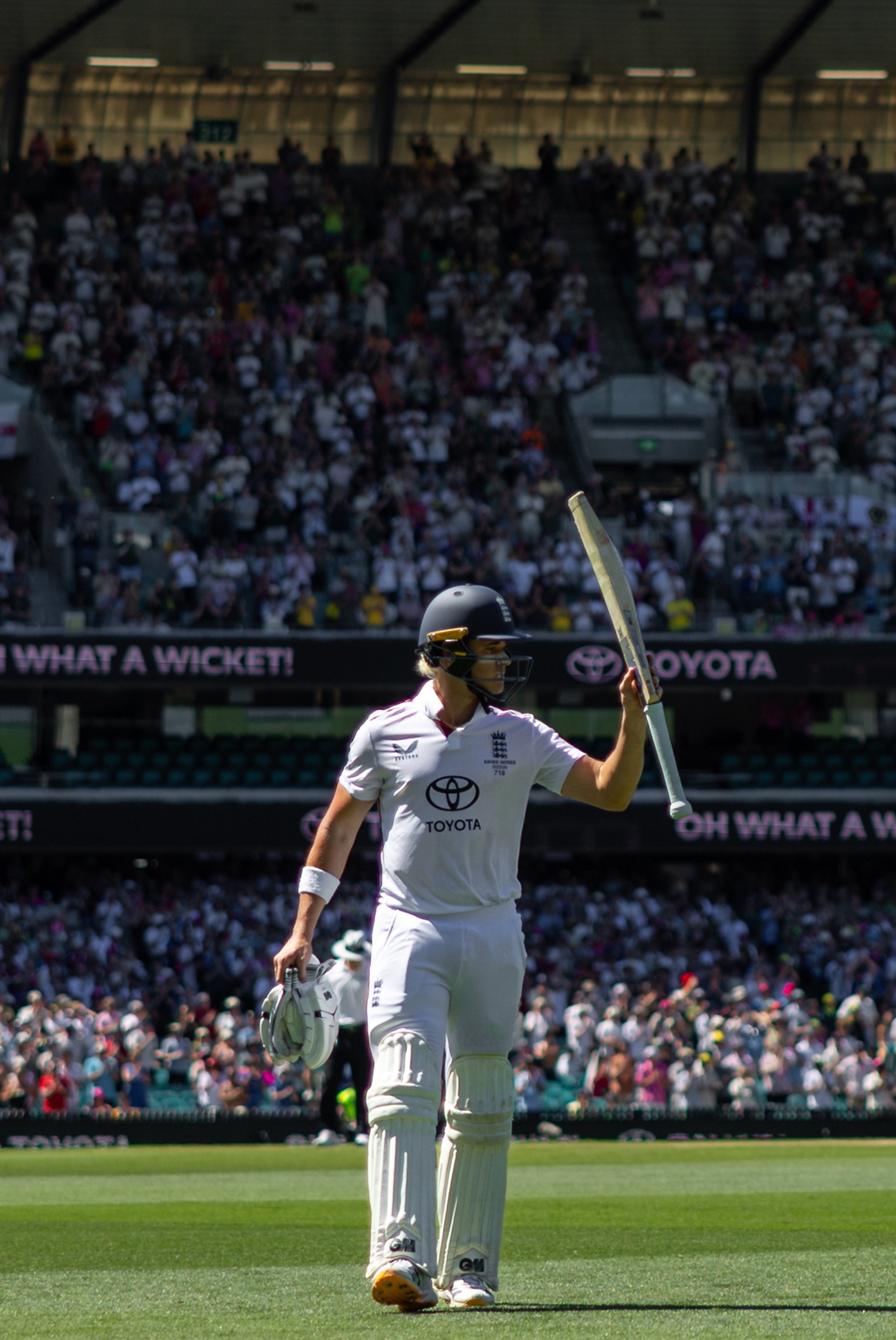 Um jogador de críquete sai do campo segurando seu taco bem alto enquanto a multidão o aplaude
