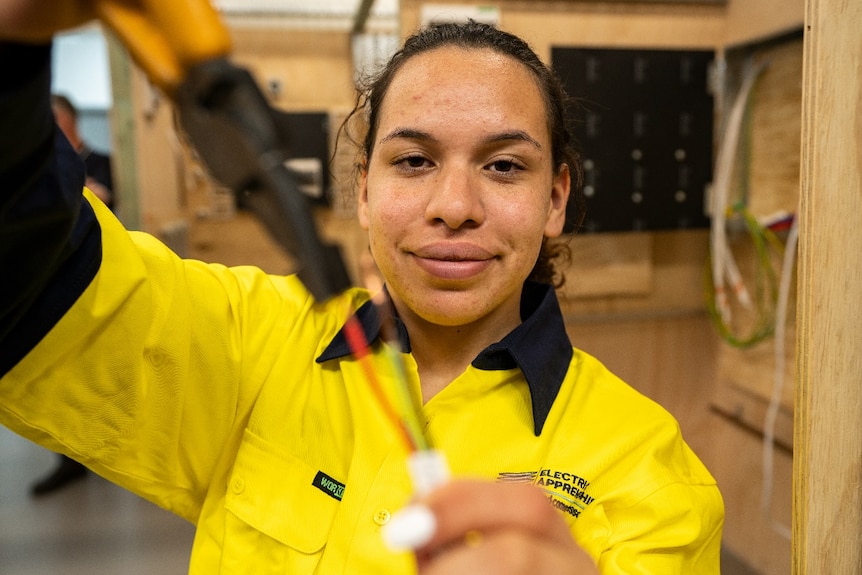 A young dark-skinned woman smiles as she holds some electrical wires with pliers.