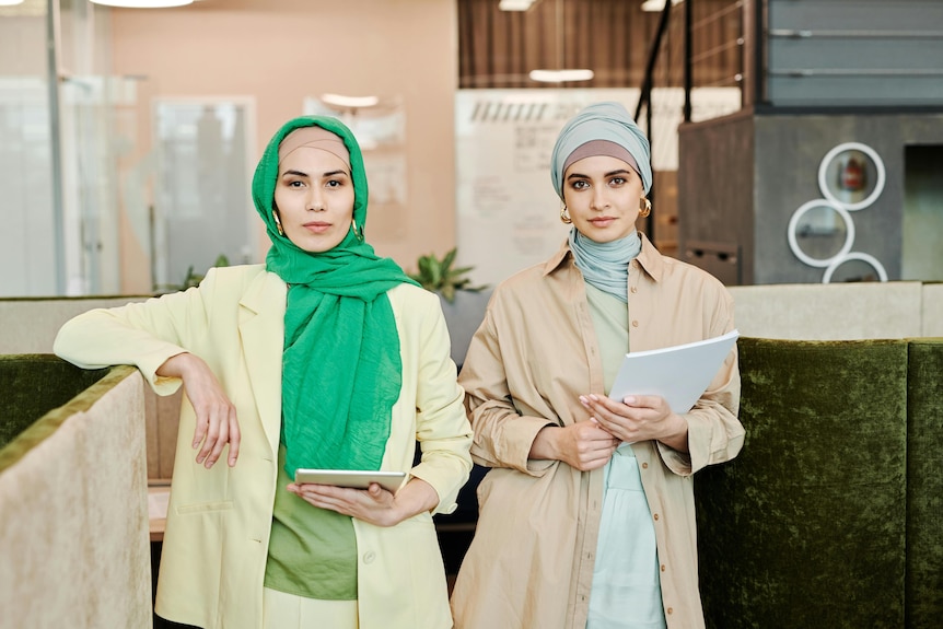 Two stylish business women looking at the camera in an office setting. Each wears a headscarf.