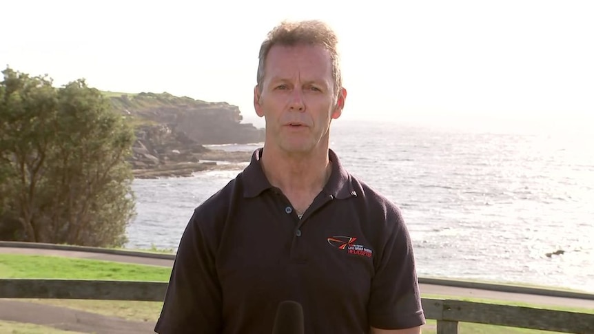 A man stands in front of the ocean with a rocky outcrop behind him.