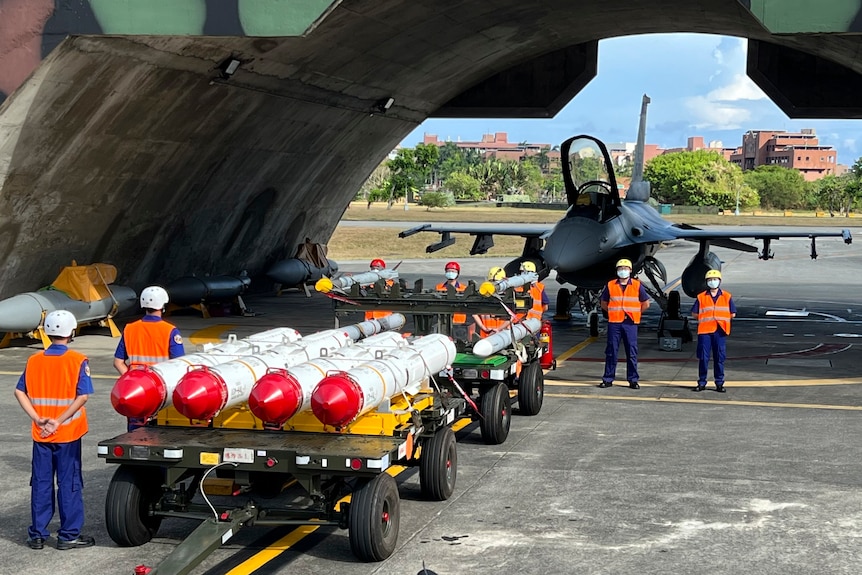 Military personnel stand beside anti-ship missiles and a fighter jet.