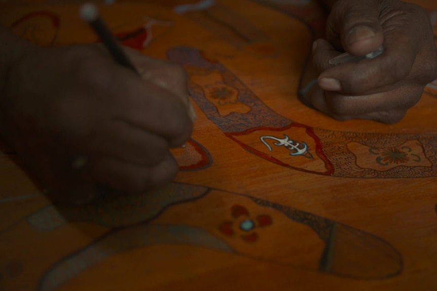 Two hands move across a wooden board with boomerangs etched and coloured onto it.