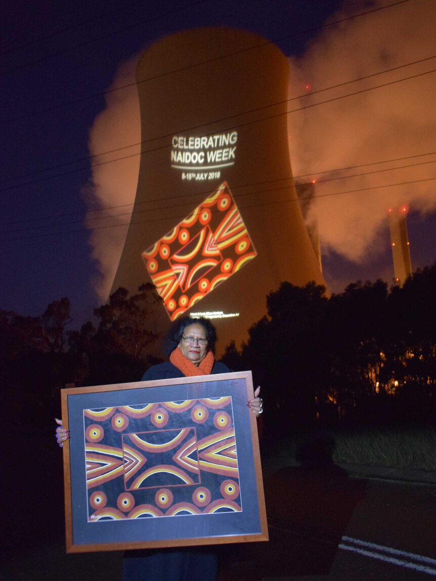 A woman holds up a painting while behind her the same image is projected much bigger in light on a power station tower.