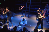 Indigenous actors on stage, dressed in black, holding boomerangs and cricket bats. A man dressed in white sits in the middle.