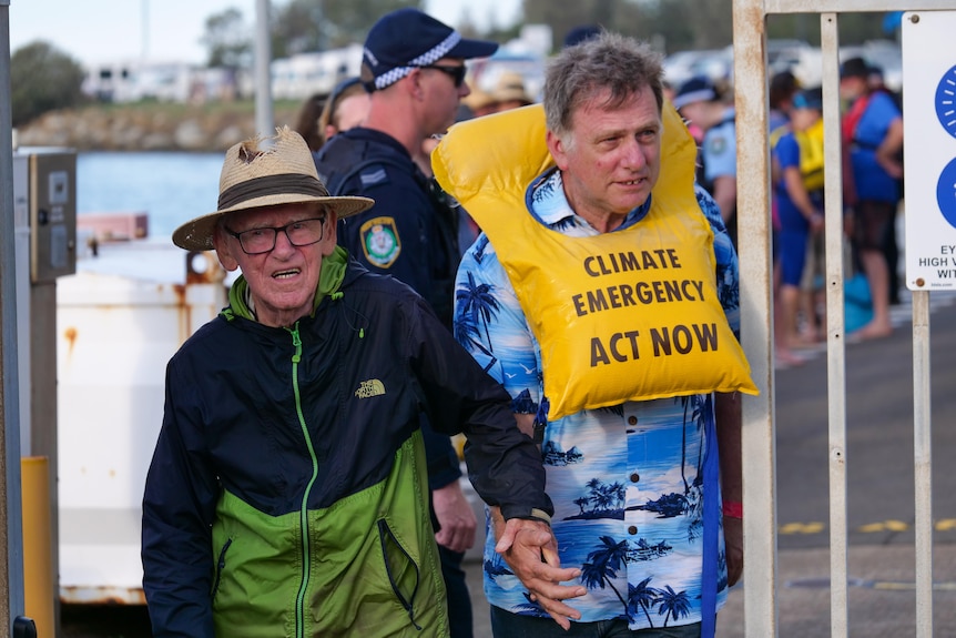 Man wearing straw hat holds the hand of man in tropical shirt wearing life jacket that says 'climate emergency act now'. 