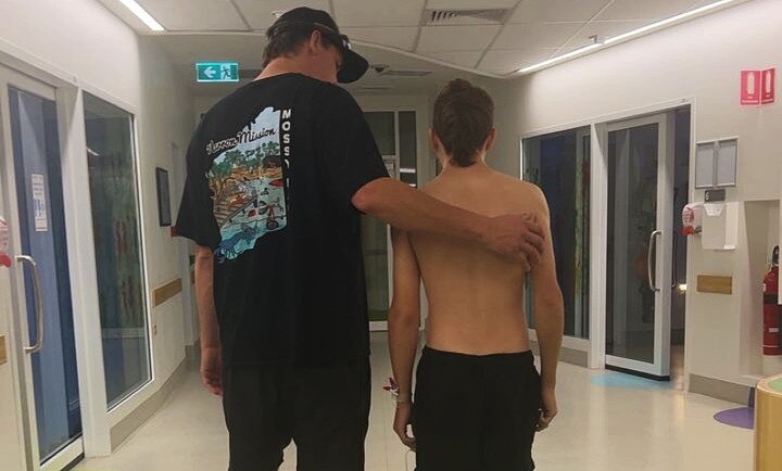 A man puts his arm around a shirtless boy as they walk down a hospital corridor.