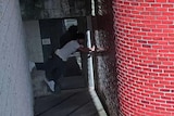 A man in a dark corner putting his hands on a red brick wall and his feet on the opposite white wall floating above ground