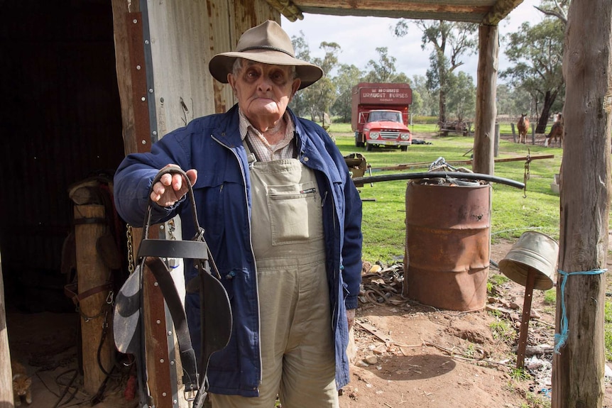 An old man standing in front of a farm shed with a truck and horses in the background, holding an old harness
