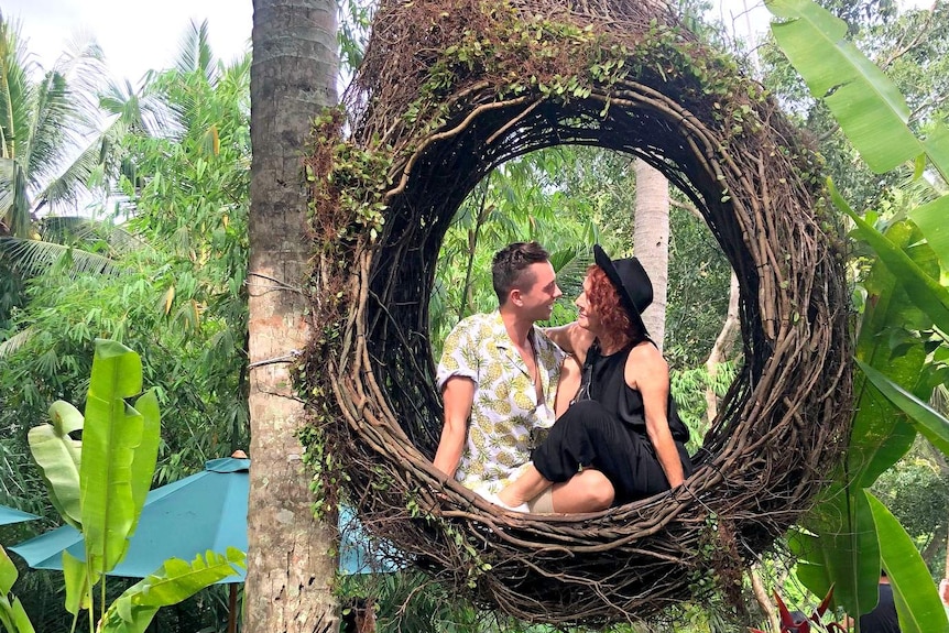 A man and a woman sit in a circular hanging seat made from woven vines or branches. Forest and shade umbrellas in background.