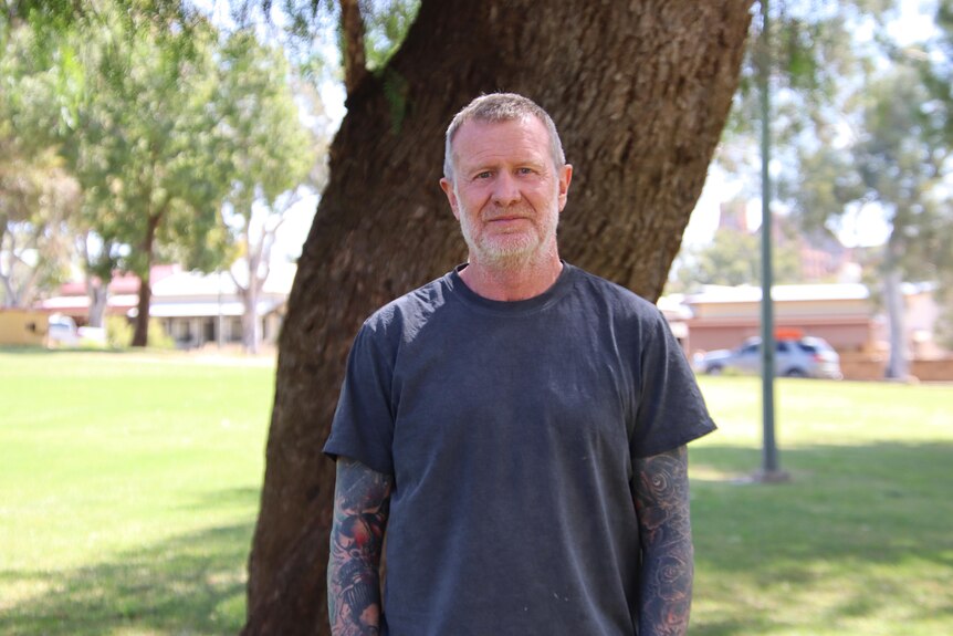 A man with a grey beard and tattoo sleeves wearing a blue t-shirt standing in front of a tree in a park