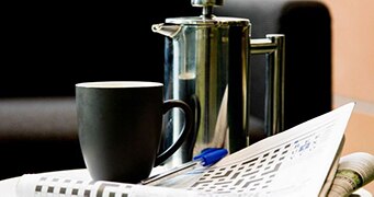 The crossword page of a newspaper lays open with a pen on it, next to a mug and coffee plunger.