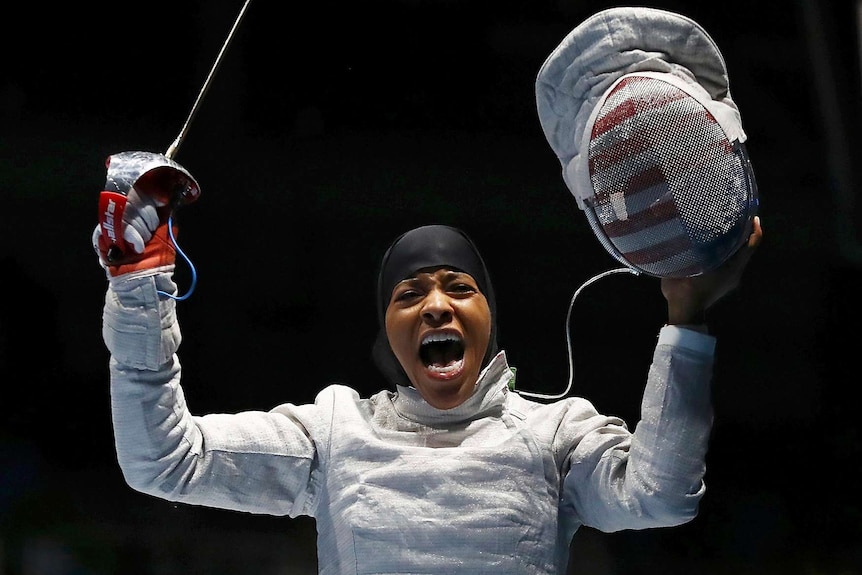 Woman in hijab and wearing fencing costume appears exultant.