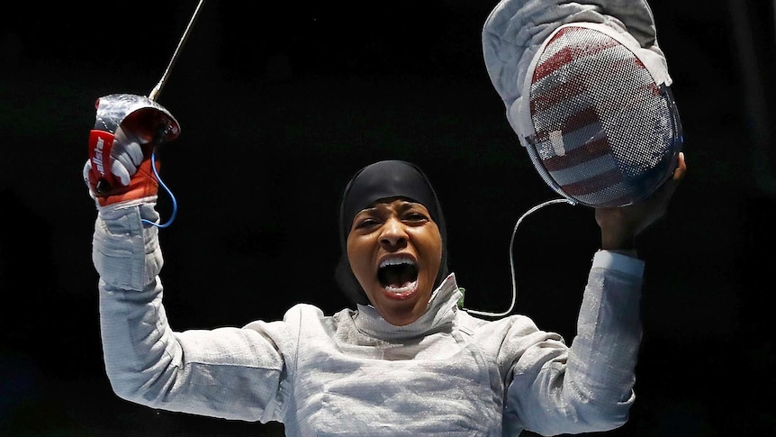Woman in hijab and wearing fencing costume appears exultant.