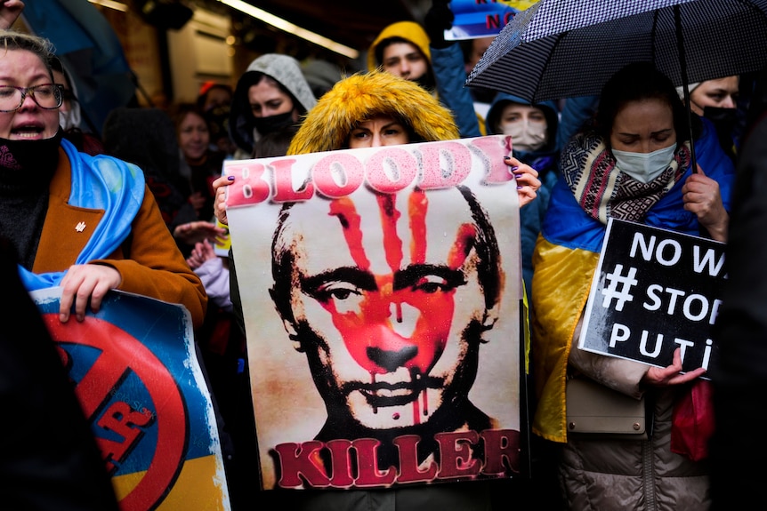 Among protesters, a woman holds a poster of Vladimir Putin with blood on his face and the words 'Bloody Killer' on it
