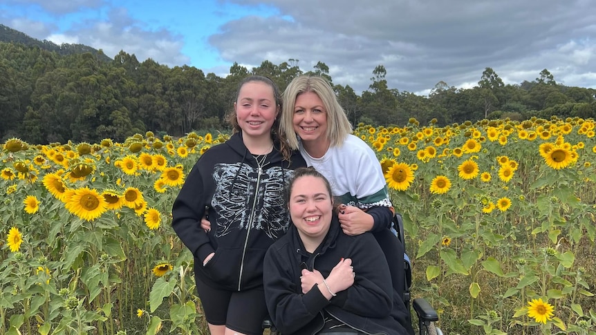 A blonde woman is hugging her two daughters in a field of sunflowers. One daughter is standing, one is using a wheelchair.