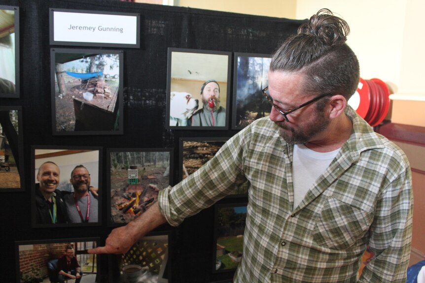 A man in a checkered shirt with a bun points to photos of him on a black wall. 