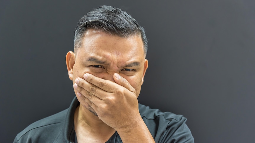 Close-up photo of man covering his nose and face with his hand and screwing up his face as if smelling something terrible.