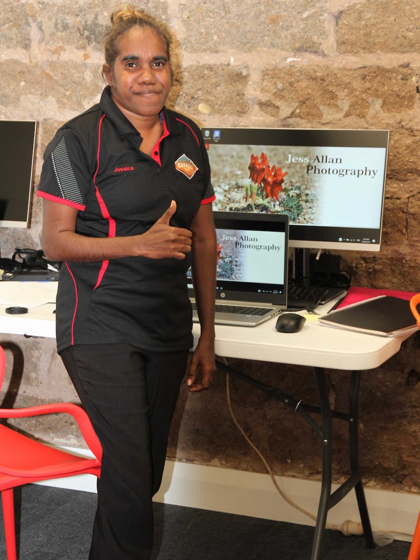 A woman gives the thumbs up as she stands in front of a computer.