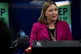 A woman stands at a podium in front of 'keep on top of COVID' signage.