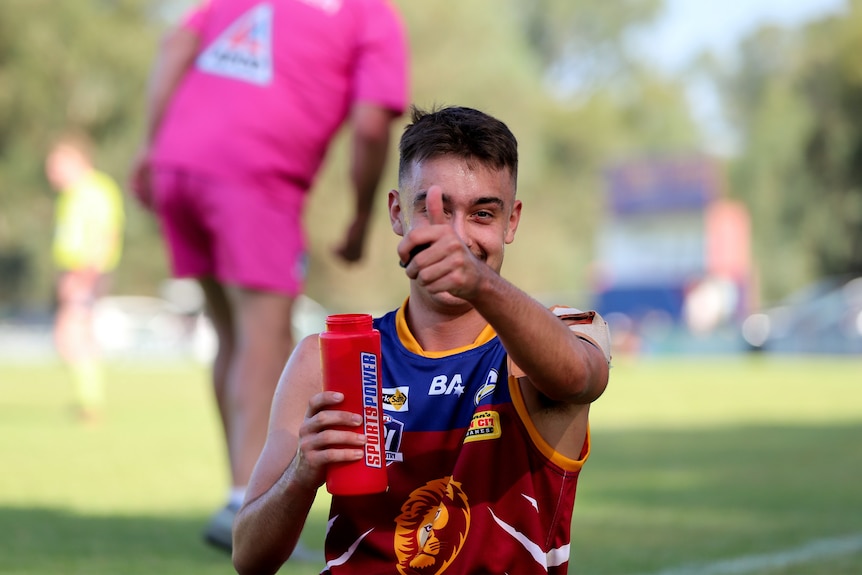 Young footballer on sidelines of green grass football oval gives thumbs up gesture.