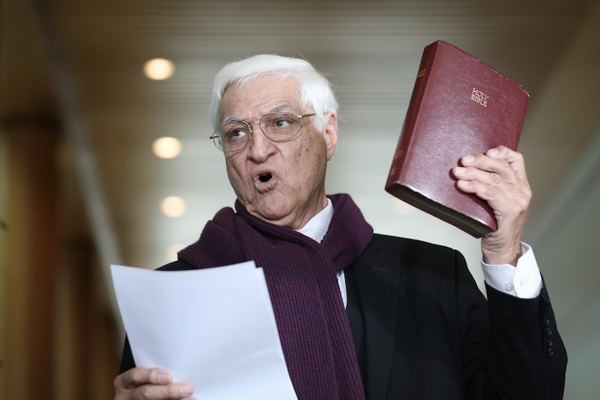 Katter sostiene una Biblia en el aire y una hoja de papel en la otra mano mientras habla.