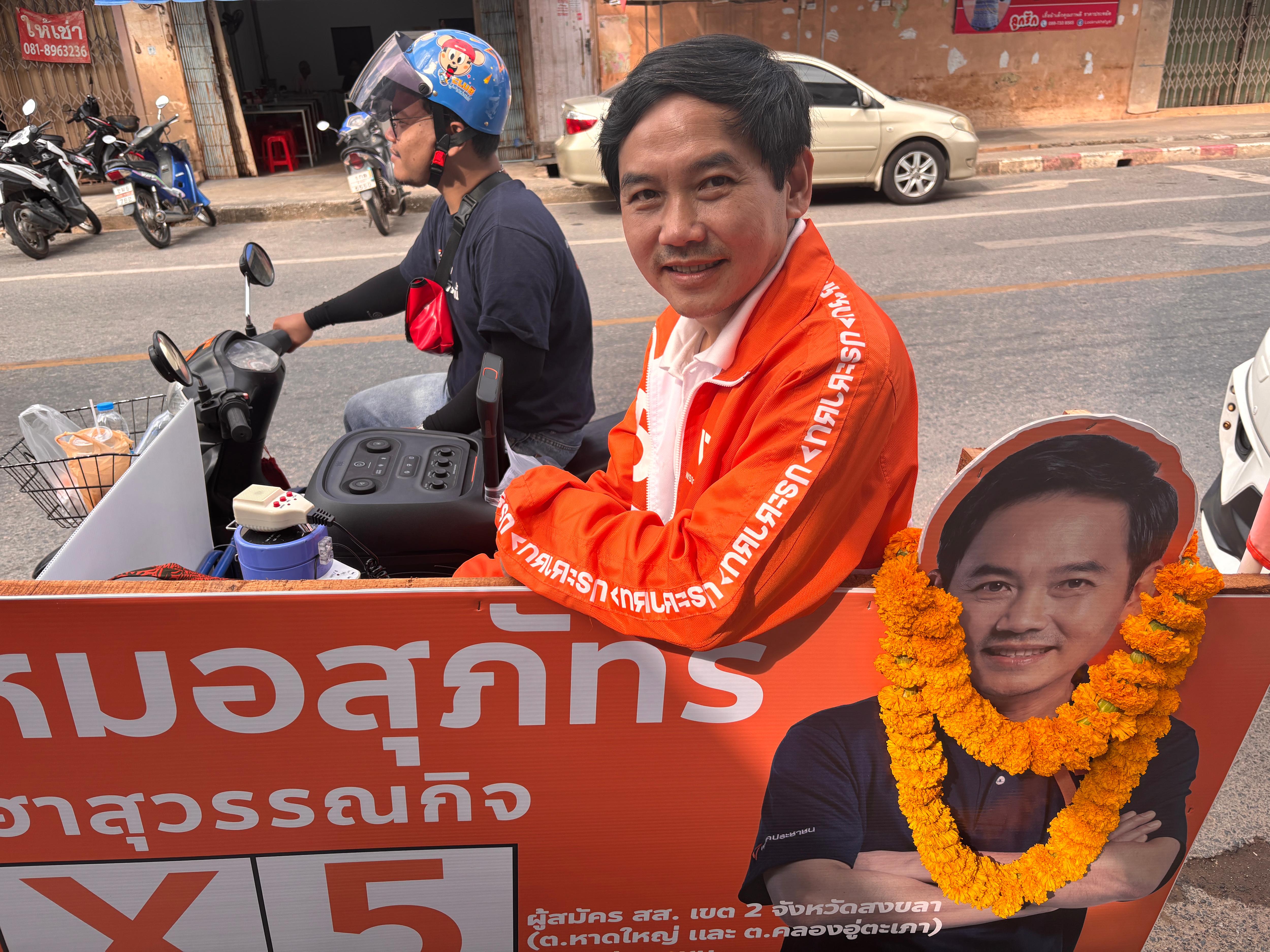 Un hombre con una chaqueta naranja brillante y un polo blanco se inclina con el rostro sobre un cartel de campaña naranja.