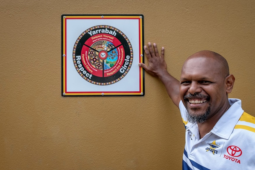 Aboriginal man wearing NRL Cowboys shirt posing next to a diagram on the wall with the words 'Yarrabah Respect Circle'.