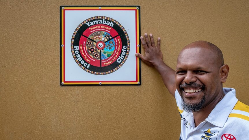 Aboriginal man wearing NRL Cowboys shirt posing next to a diagram on the wall with the words 'Yarrabah Respect Circle'.