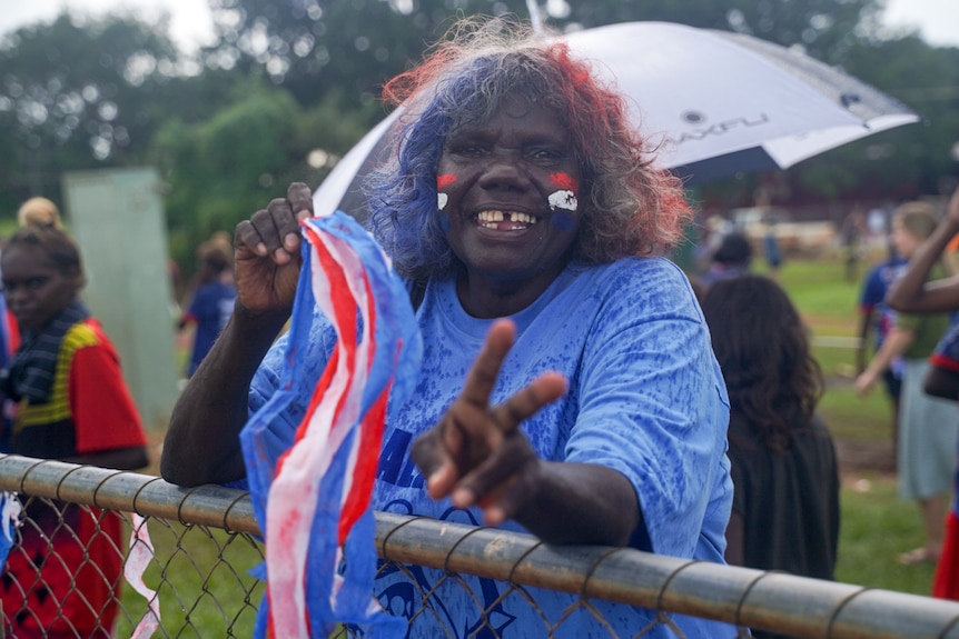 A woman at the Tiwi Islands football grand final, wearing blue with her face painted, gives a peace sign.