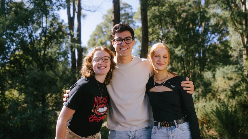 A young man has his arms around two young women as they stand in front of some trees